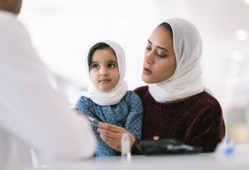 A young Muslim mother wearing a hijab takes her toddler daughter to a medical consultation. The girl is also wearing a hijab and is sitting on her mother's lap. They are seated at a table and a male doctor is seated across from them. The child is diabetic and the doctor is explaining how to use an insulin pen.
